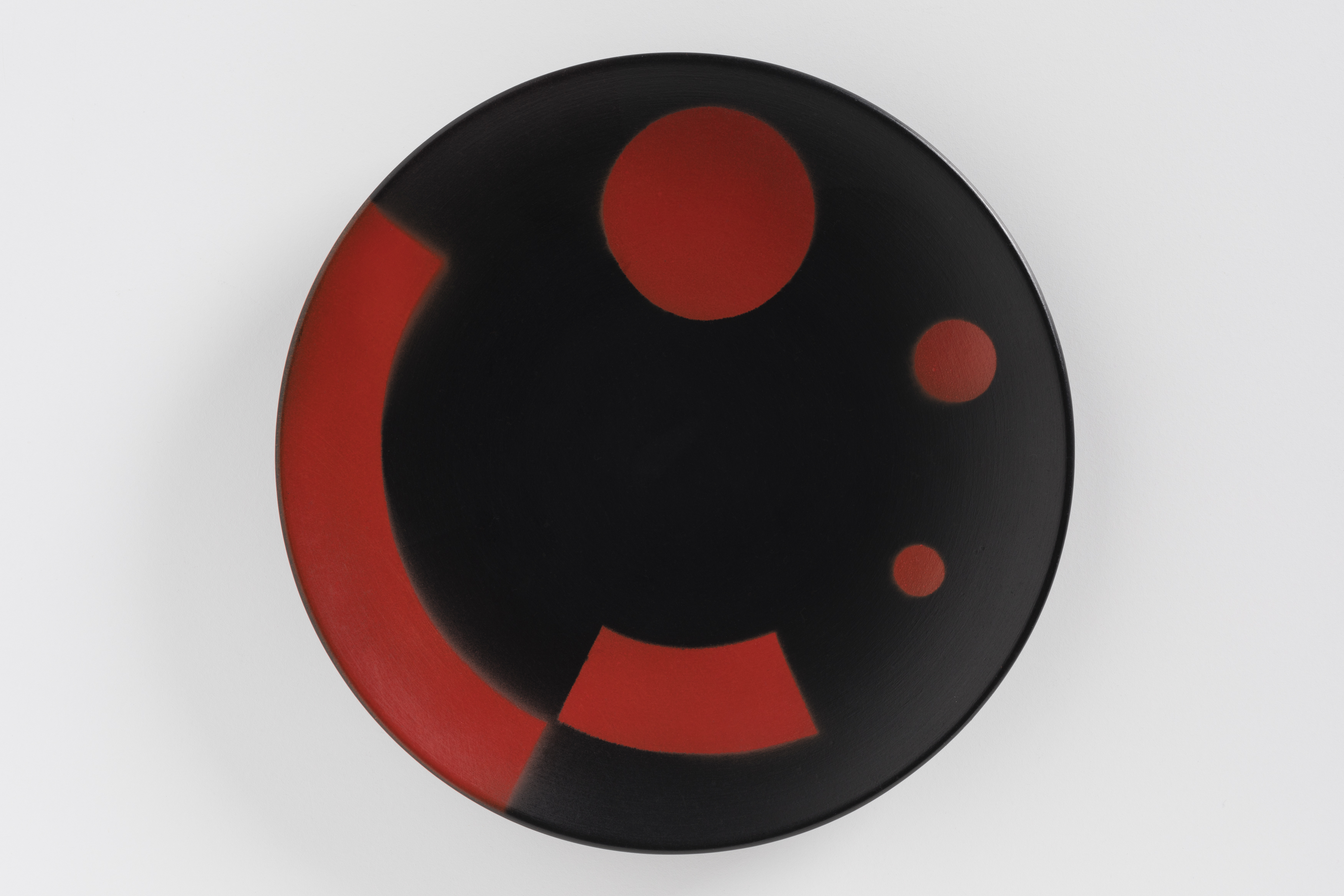A black circular plate decorated with bold red design of variously sizes dots and two curving bands.